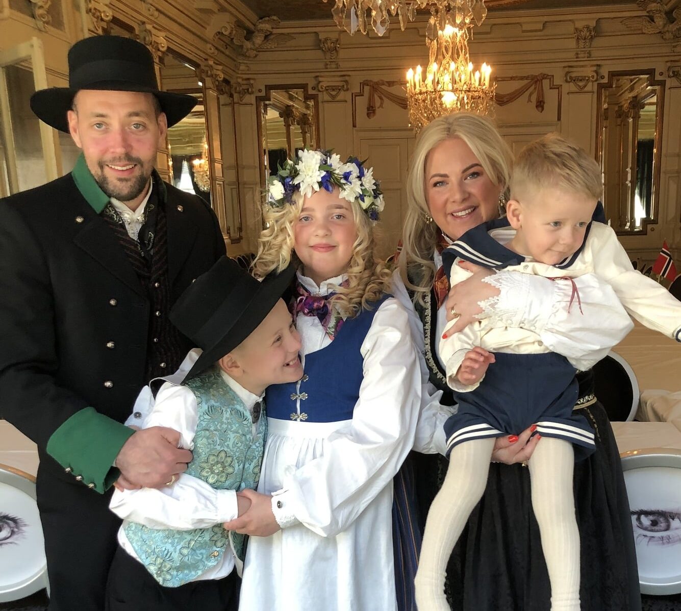 Familie på fem kledd i tradisjonelle bunader og festdrakter står samlet i et rikt dekorert rom med lysekroner og speil. To voksne og tre barn poserer tett sammen og smiler mot kamera.