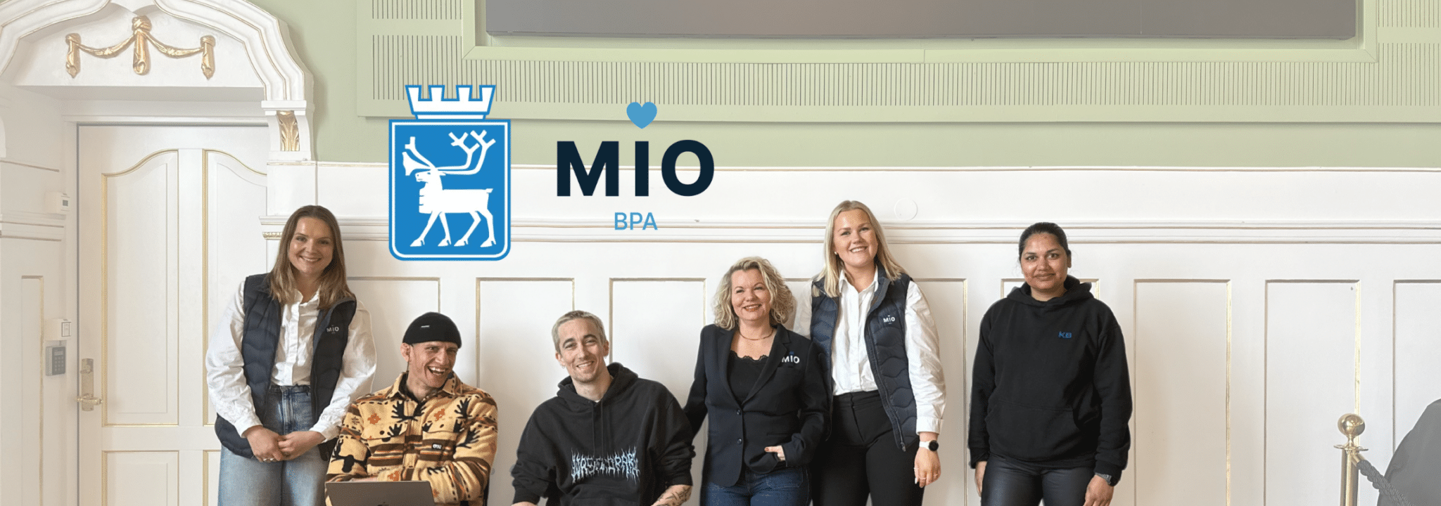 Gruppebilde av ansatte fra Mio BPA som står og sitter foran en dekorert vegg i et representativt rom. Over dem vises Mio BPA-logoen sammen med Tromsø kommune sitt kommunevåpen.