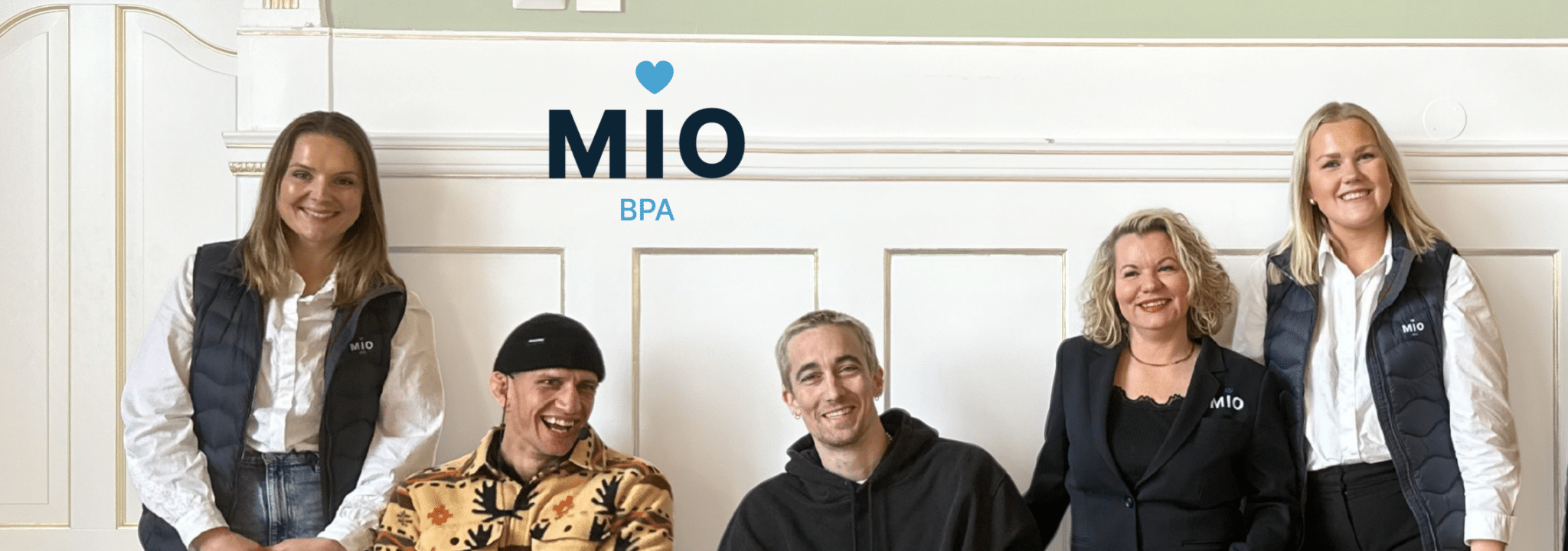 Gruppebilde av ansatte fra Mio BPA som står og sitter foran en dekorert vegg i et representativt rom. Over dem vises Mio BPA-logoen.