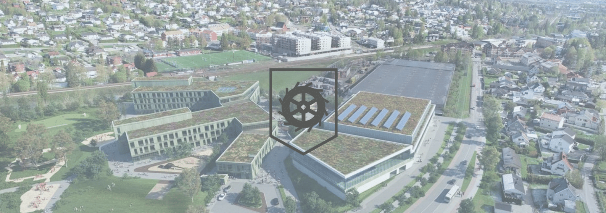 Luftfoto av et moderne bygningskompleks med grønne tak, omgitt av boligområder og veier. Midt på bildet ligger en mørk logo av Lørenskog kommune våpenskjoldet over en transparent bakgrunn.