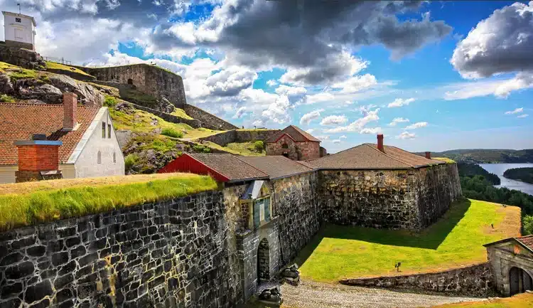 Steinfestning på en høyde med murer i flere nivåer, omgitt av trær uten blader. Et norsk flagg vaier på toppen under en blå himmel med lette skyer.