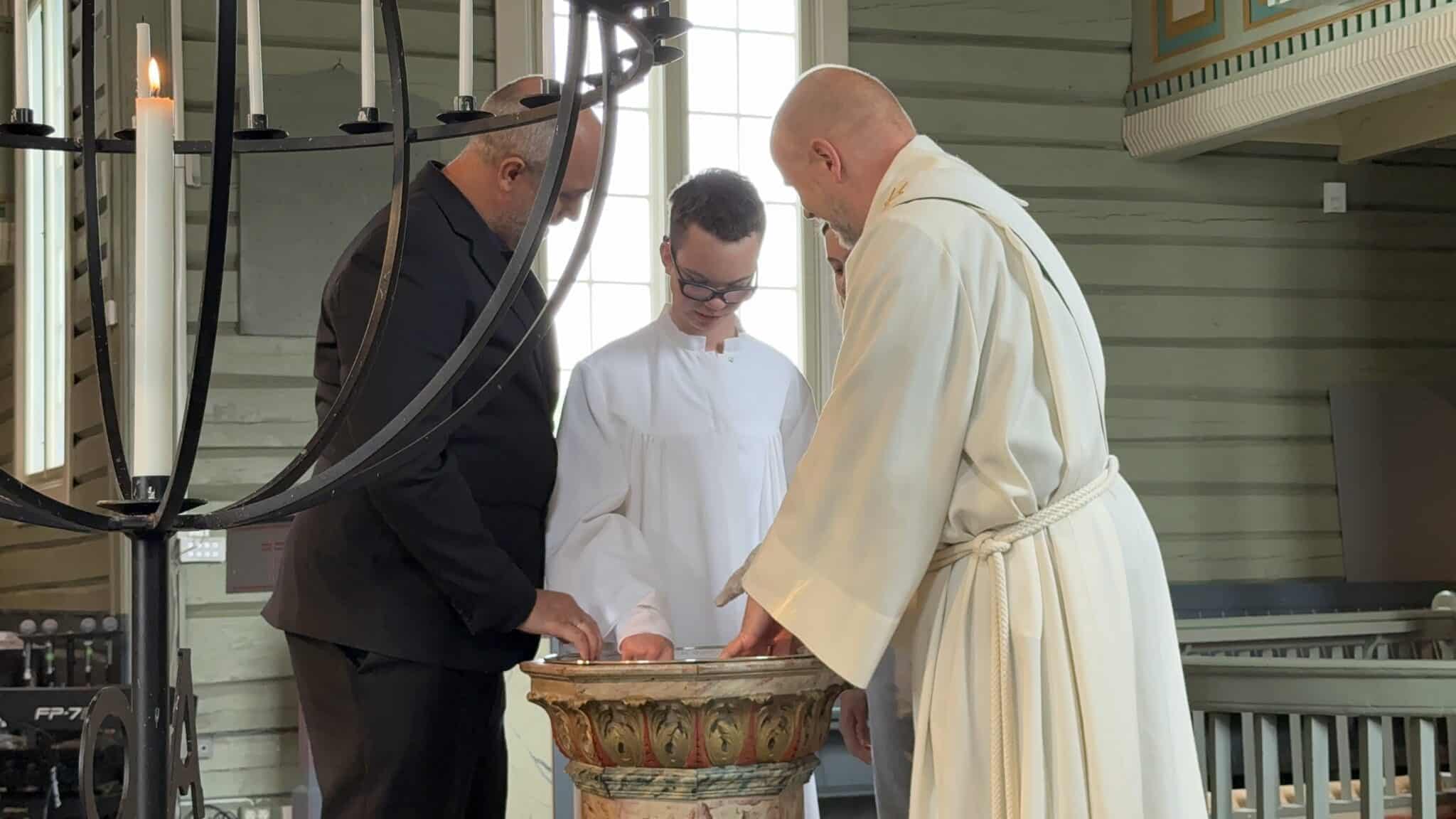 En prest i hvit messehakel, en konfirmant i hvit kappe og en voksen står samlet rundt døpefonten under en seremoni i en trekirke. Alle har hendene ved døpefonten og ser ned mot den. I forgrunnen henger en stor lysekrone med tente lys, mens dagslys strømmer inn gjennom vinduene og skaper en rolig og høytidelig stemning.