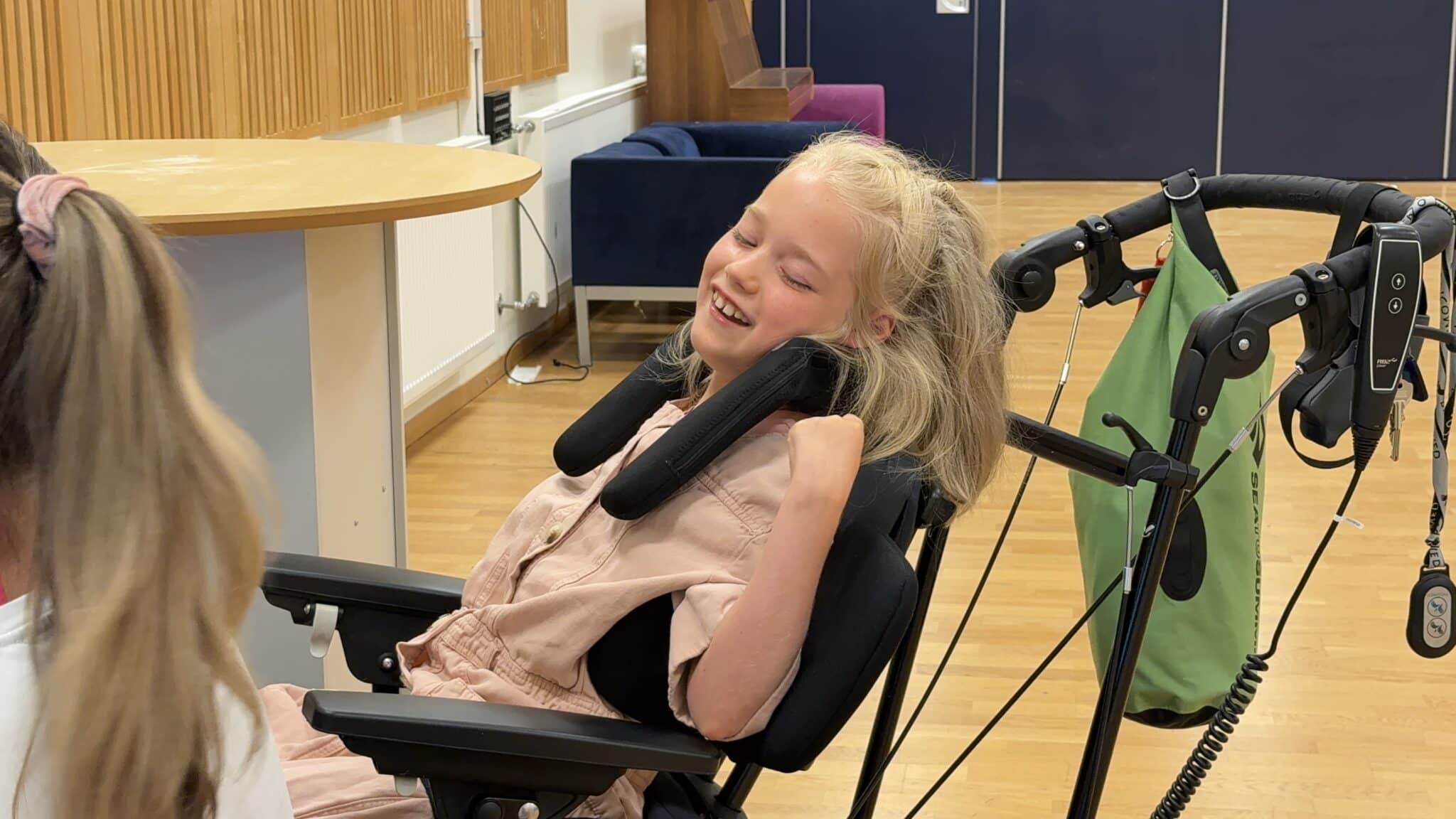 En smilende jente sitter i en elektrisk rullestol i et lyst aktivitetsrom. Hun har lyst hår, lener seg avslappet tilbake og ler mens hun ser mot en person utenfor bildet. Rommet har tregulv, bord og sitteplasser, og skaper en varm og sosial atmosfære.