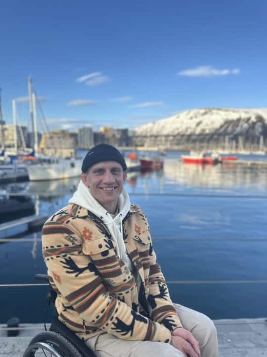 Smilende mann som sitter i rullestol ved en småbåthavn. I bakgrunnen er det seilbåter, vann med speilinger, bybebyggelse og snødekte fjell under blå himmel.