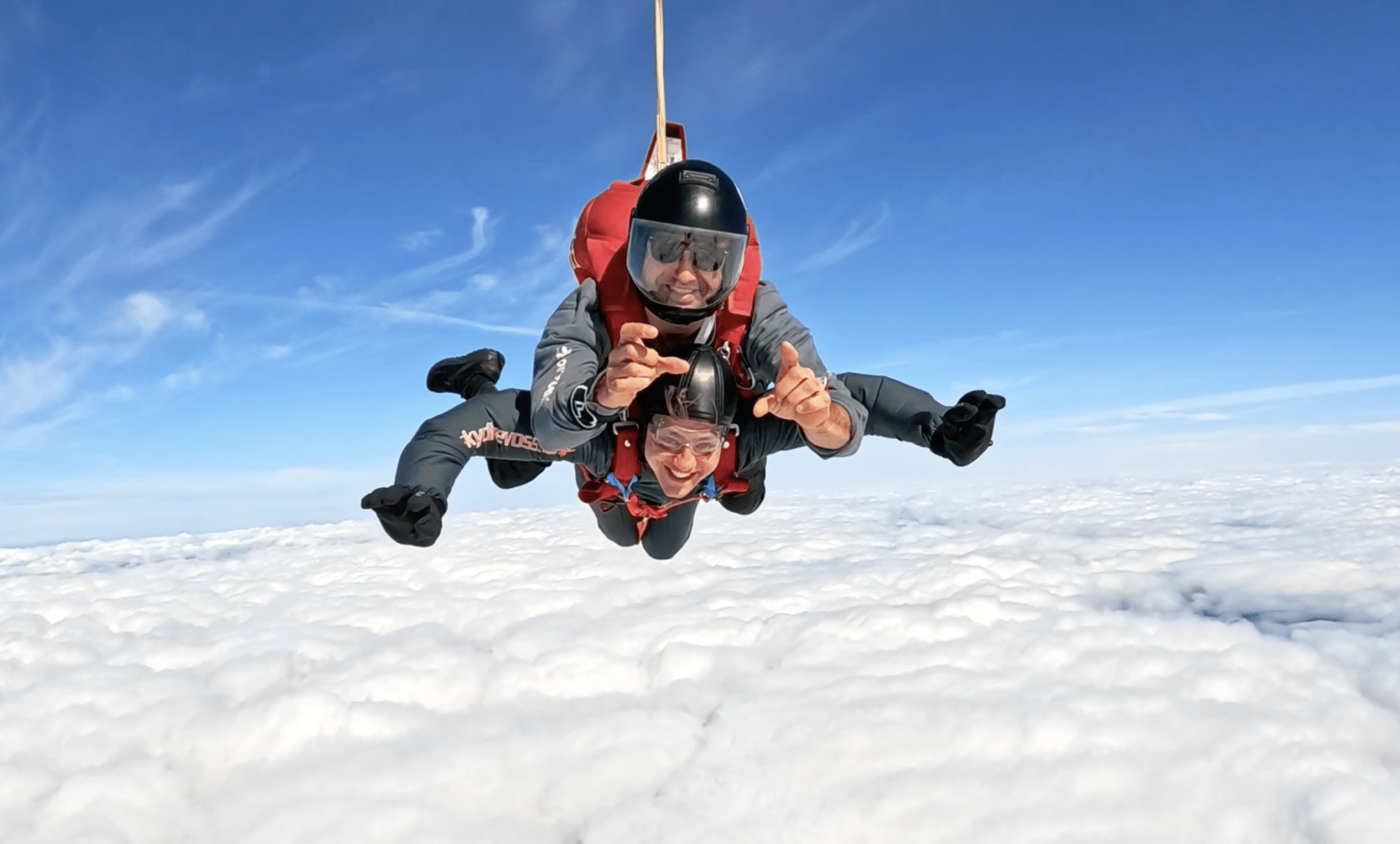 To personer i tandem fallskjermhopp svever over et hav av skyer. Den fremste personen smiler og peker mot kamera, mens instruktøren bak holder kontrollen. Begge har på fallskjermdrakter, hjelmer og vernebriller. Klar blå himmel i bakgrunnen.