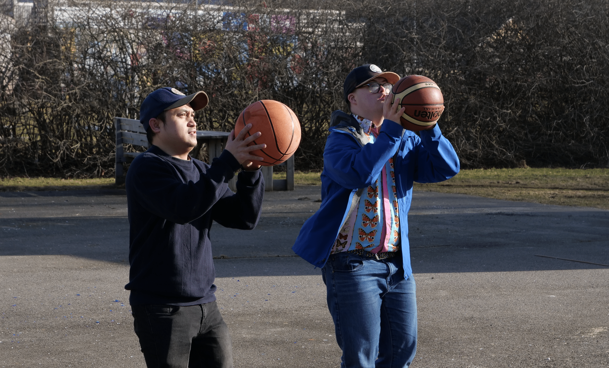 To unge menn står utendørs og holder hver sin basketball i skytestilling, klare til å kaste. De ser konsentrerte ut. Den ene har på mørk genser og caps, mens den andre har på en blå jakke og en fargerik skjorte med sommerfugler. Det er sol og bart, og en hekk vises i bakgrunnen.