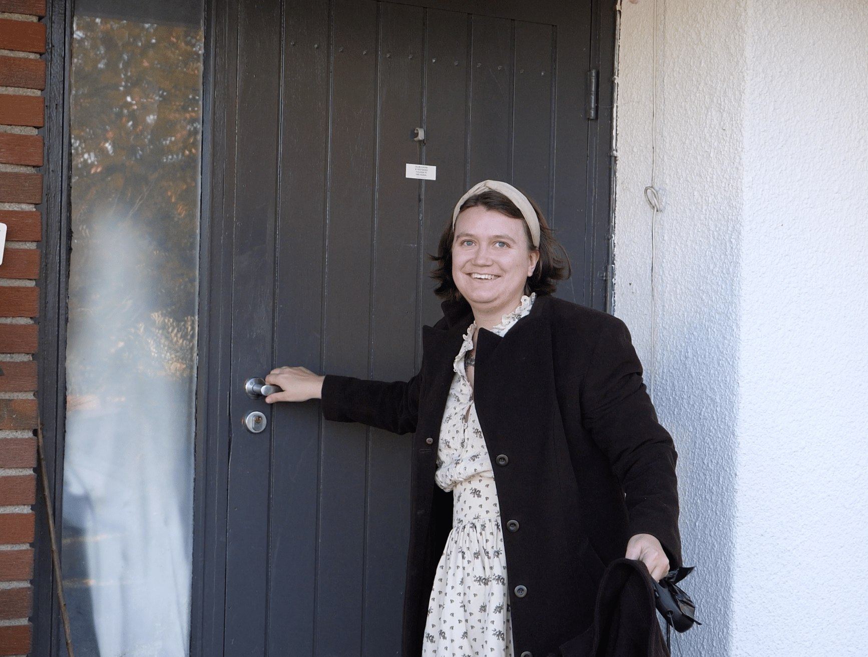 En person står foran en grå ytterdør og smiler mot kamera. Hun har en lys kjole med blomster, mørk jakke og hårbøyle, og holder i dørhåndtaket med den ene hånden og en frakk i den andre.
