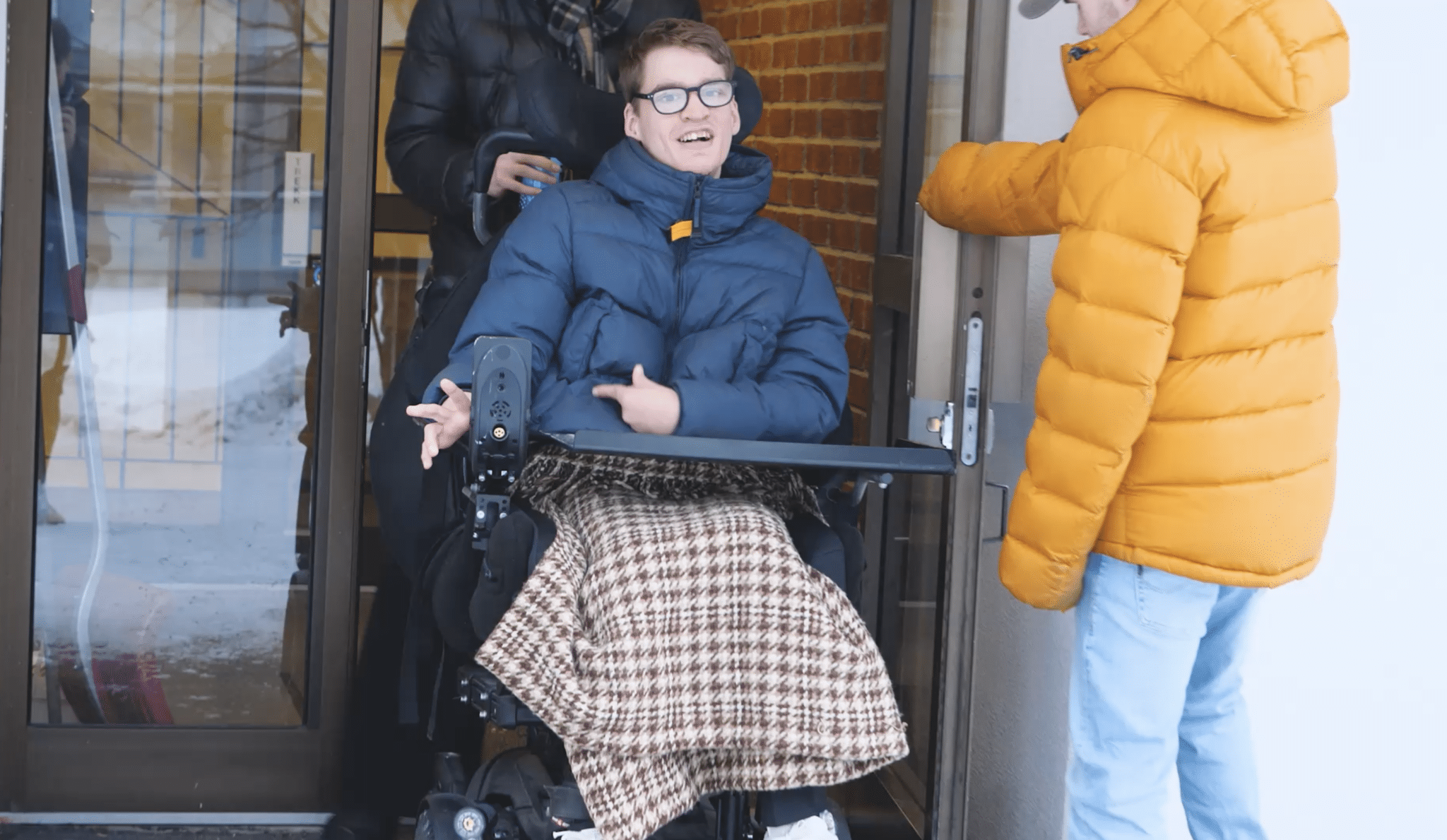 En ung mann i rullestol blir trillet ut gjennom en dør av en assistent, mens en annen person holder døren åpen. Han har på seg en blå vinterjakke og sitter med et teppe over beina, og smiler mot kamera. Bakgrunnen viser en inngang med glassdør og murvegg.