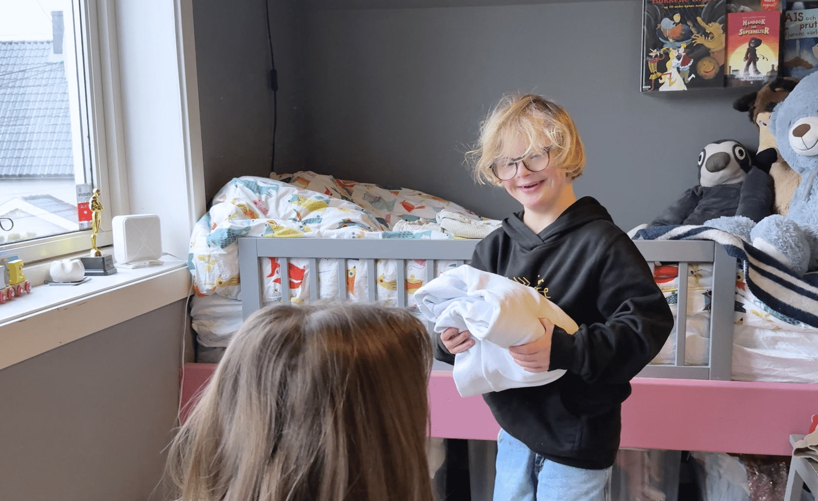 En smilende gutt med Downs syndrom står på et barnerom og holder en brettet genser, mens han ser mot en voksen person foran seg. Bak ham er en seng med fargerikt sengetøy, kosedyr og bøker, og dagslys strømmer inn fra et vindu på venstre side.