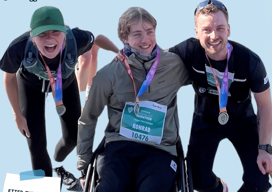 Tre personer smiler etter et løp, alle med medaljer rundt halsen. Personen i midten sitter i rullestol med startnummer “KONRAD 10476”, mens de to andre står lent inn mot ham med armene rundt skuldrene hans. Alle ser glade og stolte ut mot kamera.