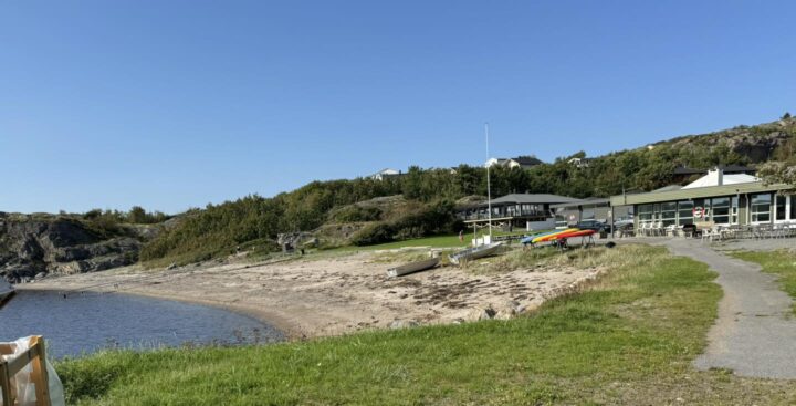 Liten sandstrand ved sjøen med robåter og kajakker på land. En sti går langs gresset mot et bygg med uteservering, omgitt av trær og svaberg under klar blå himmel.