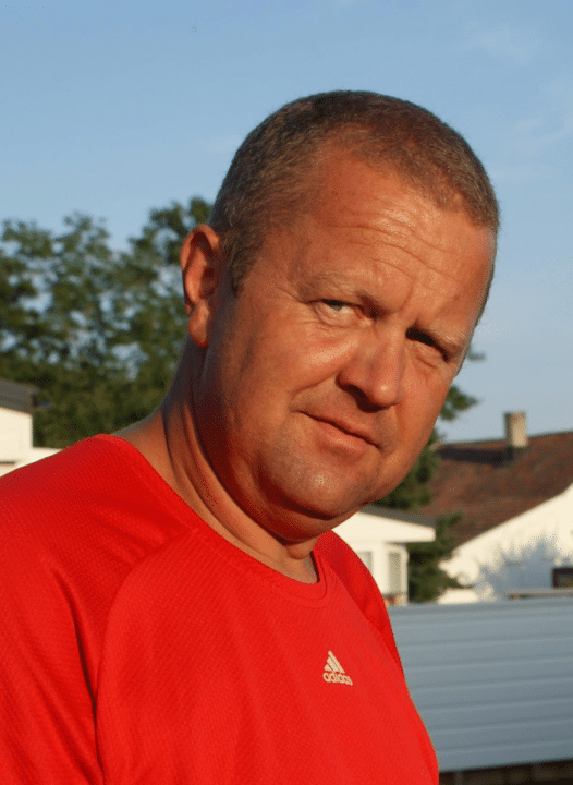 Middelaldrende mann med kort grått hår i rød Adidas-t-skjorte står utendørs i varmt kveldssollys. Han ser mot kameraet, med trær og bolighus uklart i bakgrunnen.